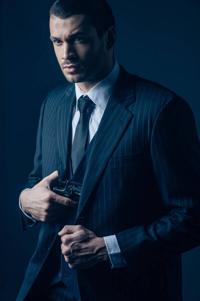 Worried and thoughtful gangster hiding weapon in suit on dark blue background