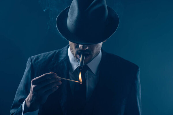 Мафиози с закрытыми глазами с войлочной шляпой освещения сигары на темно-синем фоне

