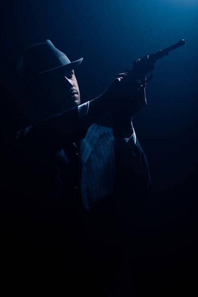 Silhouette of gangster with outstretched hands aiming revolver on dark background