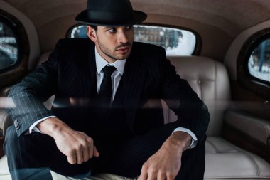 Selective focus of tense mafioso in black suit and felt hat in retro car clipart