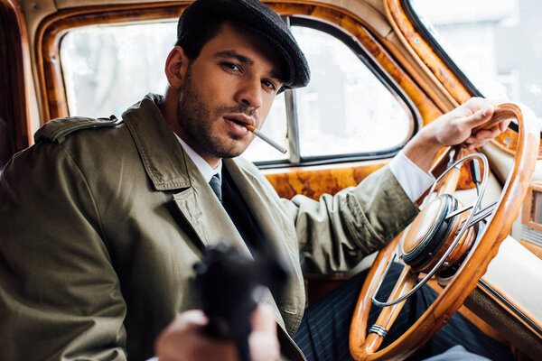 Selective focus of mafioso smoking, holding steering wheel and aiming gun in car