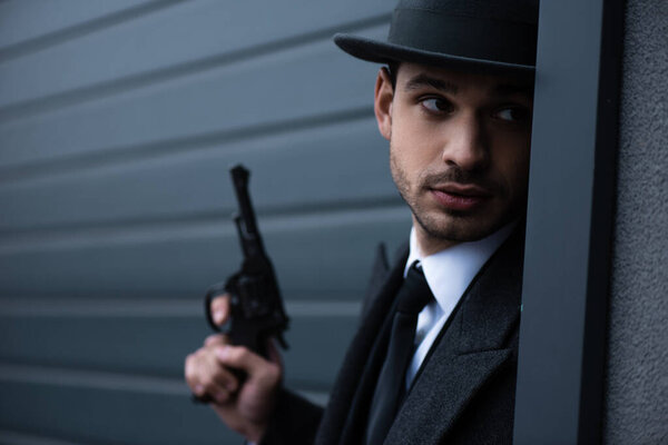 Selective focus of mafioso with revolver looking from corner near wall on street