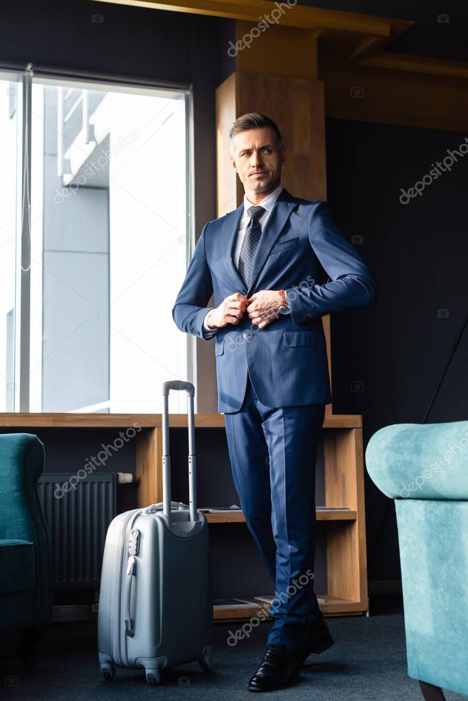 businessman in suit walking near baggage and buttoning jacket 
