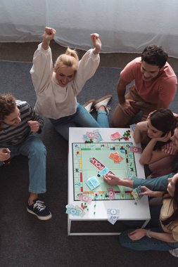 KYIV, UKRAINE - JANUARY 27, 2020: overhead view of excited girl showing winner gesture while playing monopoly game with smiling friends clipart
