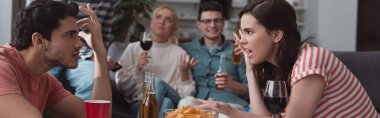 panoramic shot of girl and guy quarreling while friends sitting on sofa and drinking beverages clipart