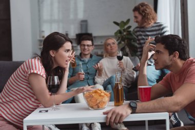 selective focus of girl and guy quarreling while friends sitting on sofa and drinking beverages clipart