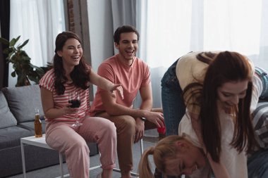 Neşeli kız elinde bir kadeh kırmızı şarap tutuyor ve evde oyun oynayan arkadaşlarını işaret ediyor.