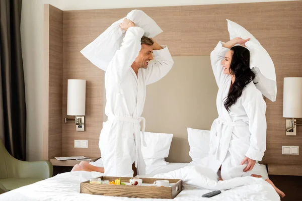 ホテルで枕と戦うバスローブの中で笑顔のボーイフレンドとガールフレンド — ストック写真