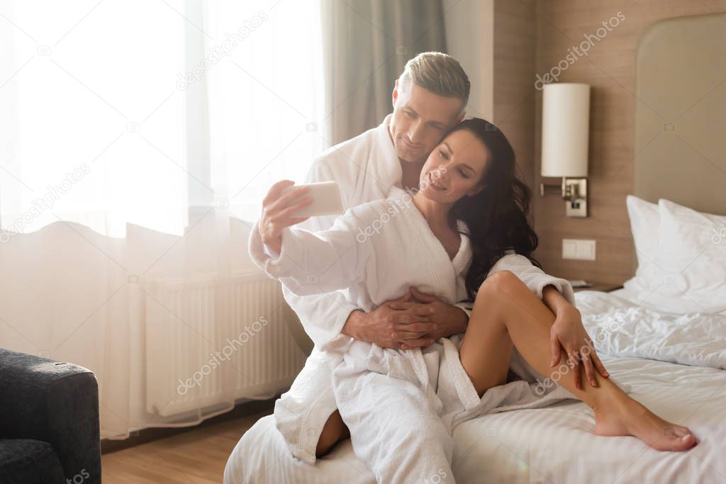 boyfriend hugging smiling girlfriend in bathrobe and she taking selfie in hotel