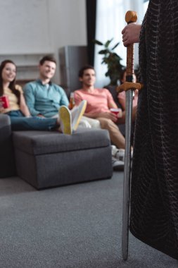 Peri kral kostümü giymiş bir adam kanepeye oturmuş gülümseyen arkadaşlarının yanında.