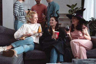 Peri kostümlü neşeli kızların yanında durup kanepede içki içen erkeklerin görüntüsü.