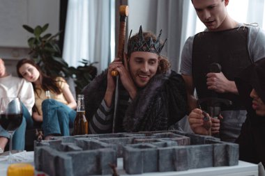 KYIV, UKRAINE - JANUARY 27, 2020: selective focus of smiling friends in fairy costumes playing labyrinth board game while bored girls sitting on sofa clipart