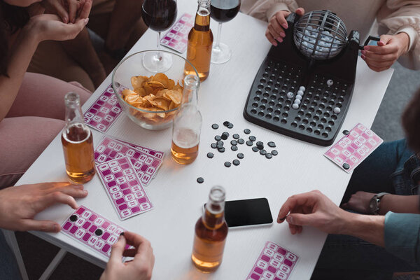 KYIV, UKRAINE - JANUARY 27, 2020: cropped view of friends sitting at table with lotto balls dispenser, cards, covering chips and smartphone with blank screen