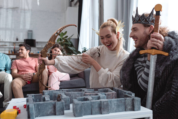 KYIV, UKRAINE - JANUARY 27, 2020: cheerful girl and guy in fairy costumes playing labyrinth game near bored friends sitting on sofa