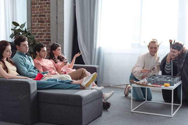 KYIV, UKRAINE - JANUARY 27, 2020: cheerful girl and guy in fairy costumes playing labyrinth game near bored friends sitting on sofa