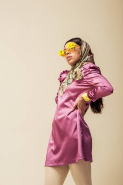 Güneş gözlüklü, çiçekli, eşarplı ve elbisesi bej renkli kalçasında duran modaya uyan Afrikalı Amerikalı kız. 