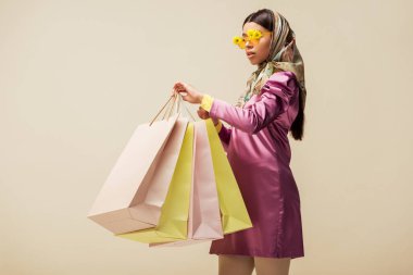 stylish african american girl in sunglasses with flowers, head scarf and dress holding shopping bags isolated on beige  clipart