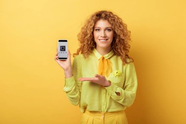 Kyiv, Ukrayna - 4 Şubat 2020: Sarı ekranda izole edilmiş süper uygulamalı akıllı telefonu işaret eden mutlu kızıl kadın 