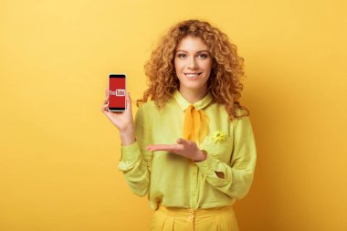 Kyiv, Ukrayna - 4 Şubat 2020: Sarı ekranda izole edilmiş youtube uygulaması ile akıllı telefonu gösteren mutlu kızıl kadın 