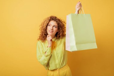 pensive redhead woman holding shopping bags on yellow  clipart