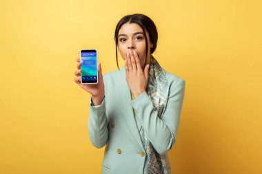 Şaşırmış Afrikalı Amerikalı kız yüzünü örtüyor ve elinde sarı telefon uygulaması tutuyor. 
