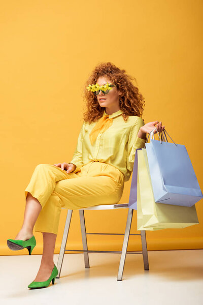 trendy redhead woman in sunglasses with flowers holding shopping bags while sitting on yellow 