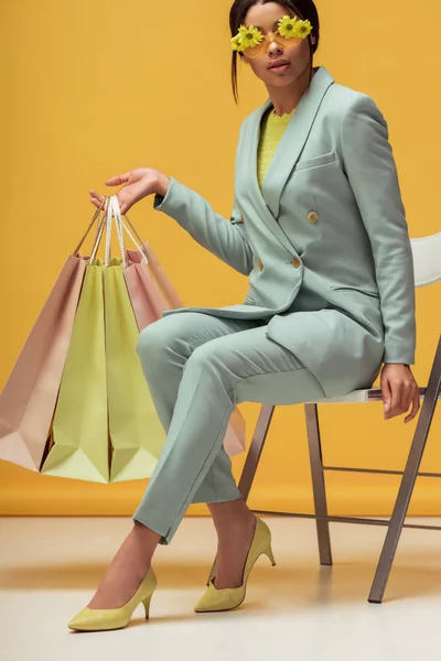 スーツ姿のアフリカ系アメリカ人女性黄色に座っながらショッピングバッグを保持花とサングラス — ストック写真