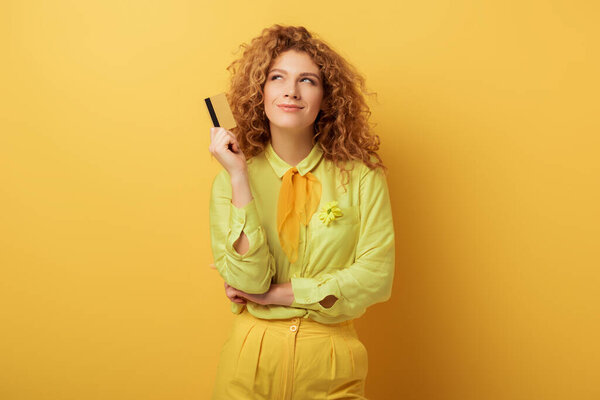 smiling redhead girl holding credit card while thinking on yellow 