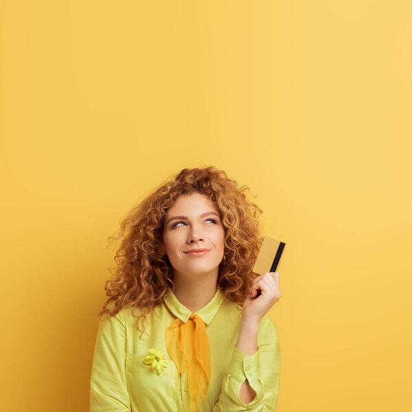 smiling redhead woman holding credit card while thinking on yellow 