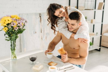 Beyaz tişörtlü seksi kız mutlu üstsüz erkek arkadaşına sarılıyor mutfakta kahvaltı yapıyor.