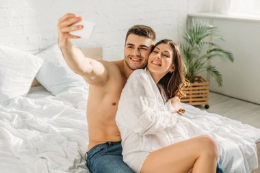 Mutlu gömleksiz adam akıllı telefondan beyaz gömlekli mutlu kız arkadaşıyla selfie çekiyor.