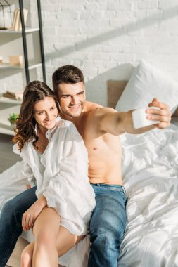 Seçici odak noktası, yatak odasında seksi kız arkadaşıyla akıllı telefondan selfie çeken neşeli bir adam.