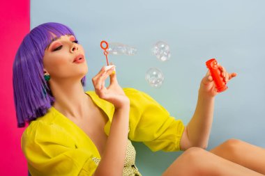 happy attractive girl in purple wig as doll blowing soap bubbles while sitting in blue box, isolated on pink clipart