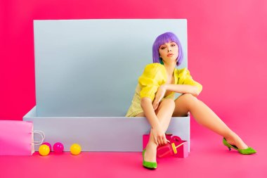 bored girl in purple wig as doll sitting in blue box with balls and shopping bags, on pink clipart