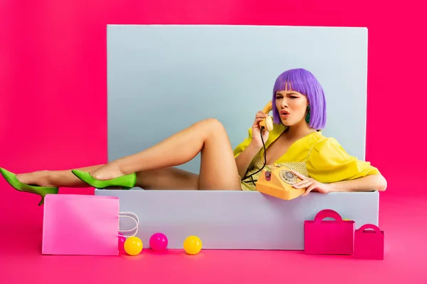 穿着紫色假发的咄咄逼人的女孩 就像一个玩具娃娃 坐在蓝色的盒子里 提着球和购物袋 坐在粉色的老式手机上聊天 — 图库照片