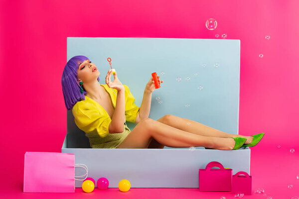 attractive girl in purple wig as doll blowing soap bubbles while sitting in blue box with balls and shopping bags, on pink