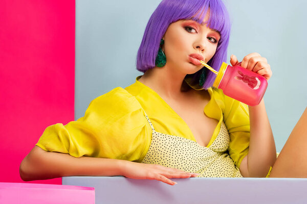 beautiful girl in purple wig as doll drinking from jar while sitting in blue box, isolated on pink