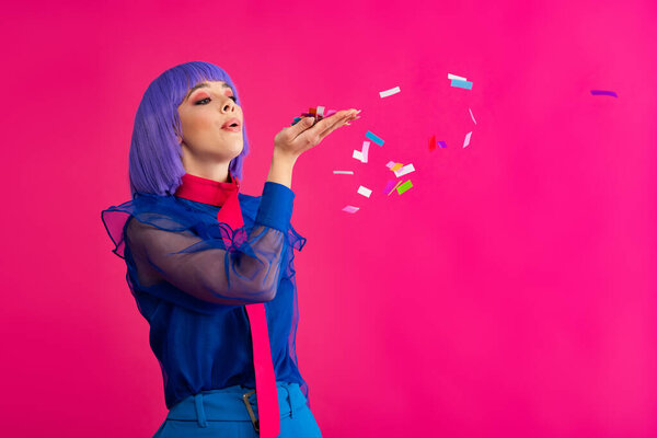 beautiful fashionable pop art girl in purple wig blowing confetti, isolated on pink
