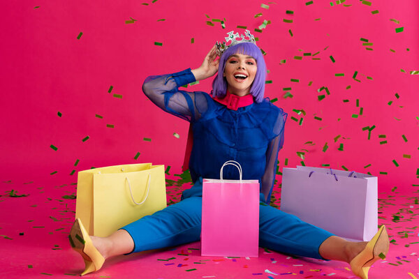 счастливая девушка в фиолетовом парике и короне сидя с пакетами покупок и праздничные конфетти, на розовый
