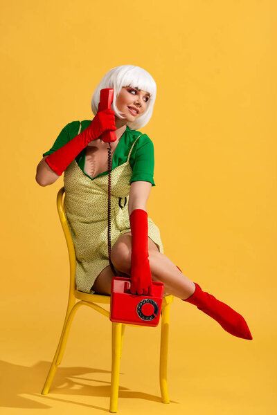 smiling girl in white wig talking on retro phone while sitting on chair, on yellow