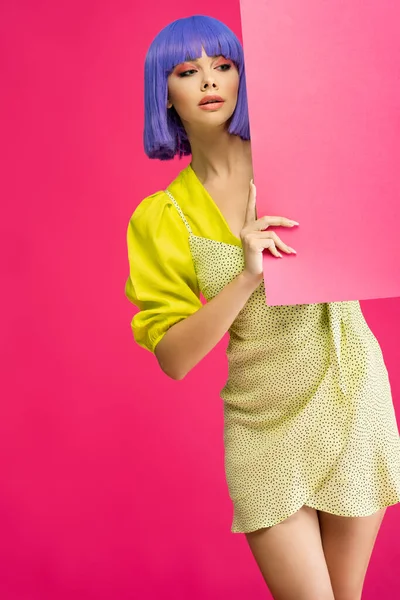 身穿紫色假发和黄色连衣裙的漂亮女孩 手持空白标语牌 与粉色隔离 — 图库照片