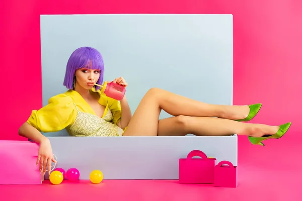 Bored girl in purple wig as doll drinking from jar while sitting in blue box with balls and shopping bags, on pink — Stock Image Bored Girl Purple Wig Doll Drinking Jar While Sitting Blue Stock Image