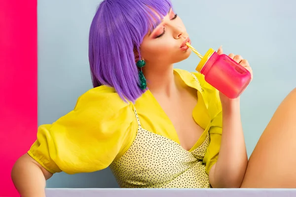 Beautiful pop art girl in purple wig as doll drinking from jar while sitting in blue box, isolated on pink — Stock Image Beautiful Pop Art Girl Purple Wig Doll Drinking Jar While Royalty Free Stock Photos