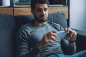 Trauriger Mann schaut Pornos auf Smartphone auf Couch im Wohnzimmer
