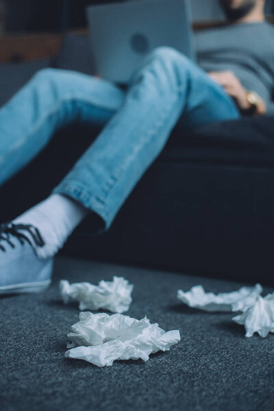 Selective focus of crumpled napkins on floor near man watching pornography on laptop on couch 