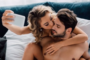 Yatakta akıllı telefonla selfie çekerken üstsüz erkek arkadaşı öpen çekici bir kadının seçici odak noktası. 