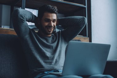 Kanepede dizüstü bilgisayar izlerken gülümseyen yakışıklı bir adam. 