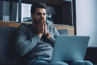 Şok olmuş bir adam, elleri ağzının kenarında oturmuş laptopta porno seyrediyor.