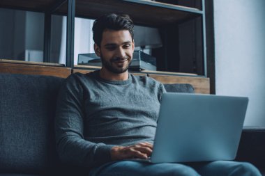 Genç adam oturma odasında dizüstü bilgisayarda porno izlerken gülümsüyor.