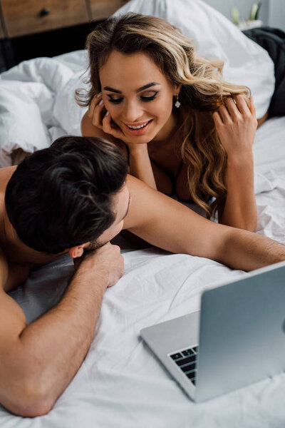Selective focus of sexy woman looking at boyfriend near laptop on bed 
